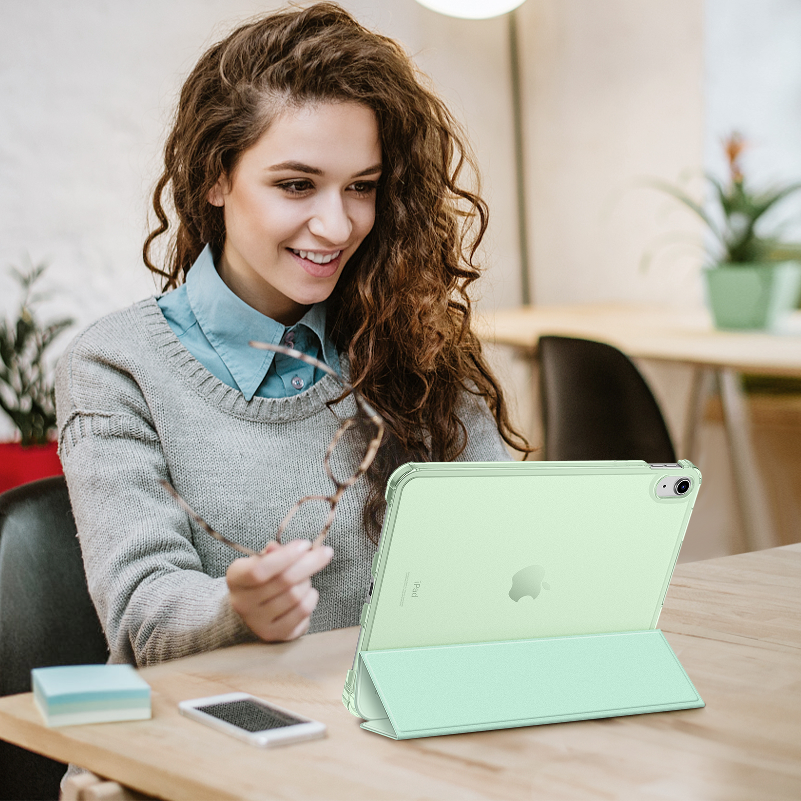 Eine Frau hält eine Brille und betrachtet ein grünes iPad in einem Gehäuse auf einem Tisch.