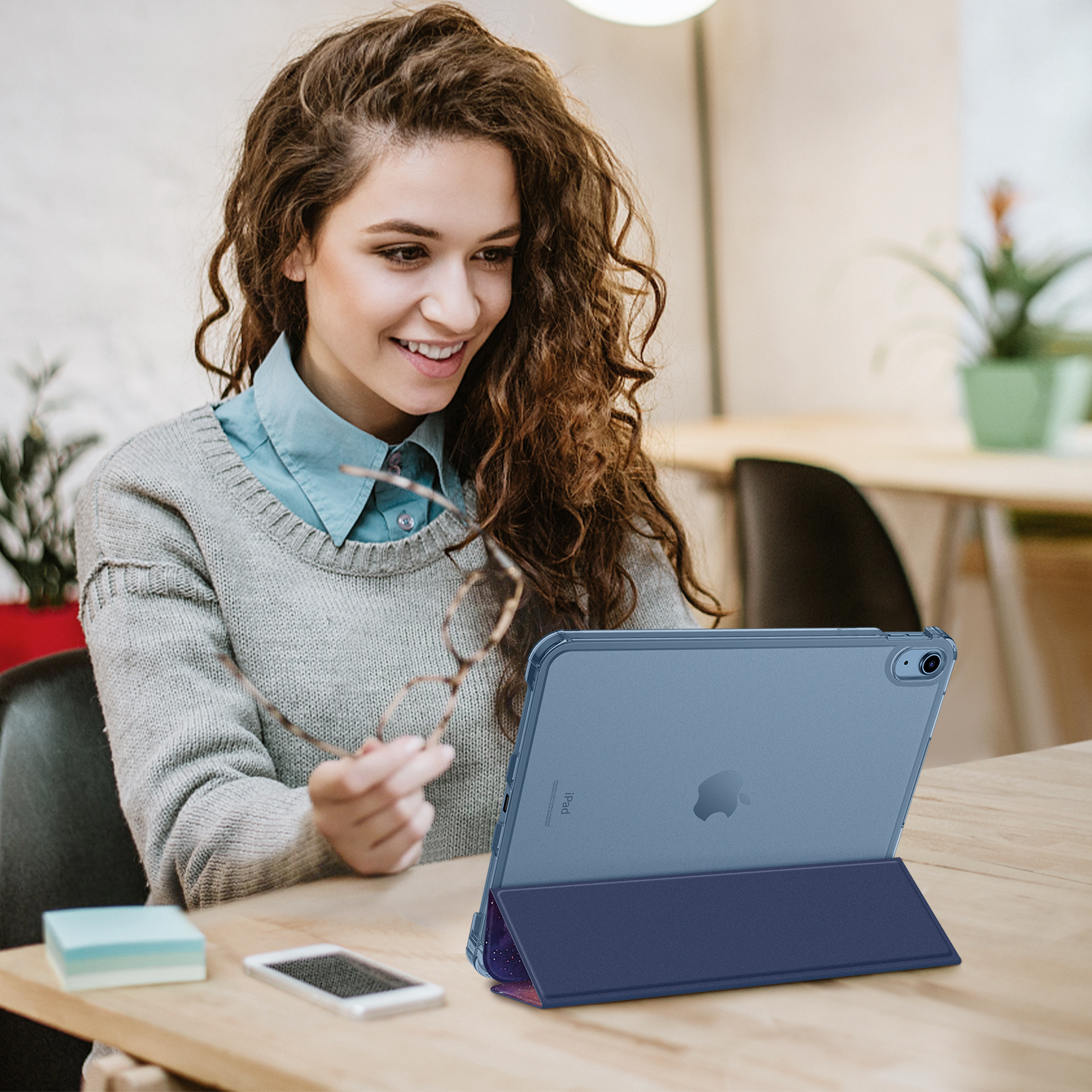 Frau hält Brille, schaut auf ein iPad mit Hülle auf einem Holztisch.