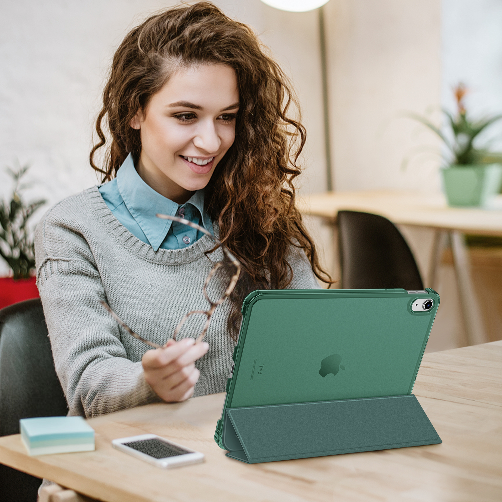Frau, die eine Brille hält, blickt auf ein grünes Tablet auf einem Holztisch.
