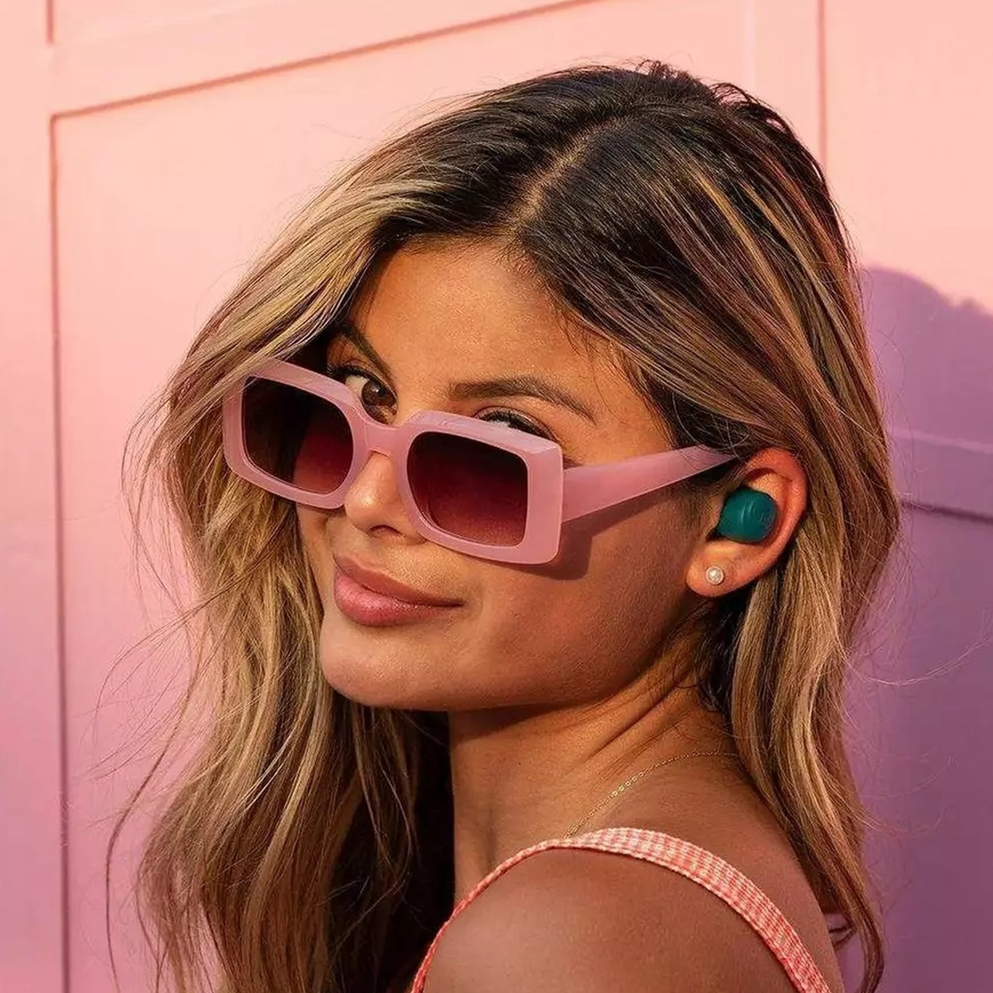 Kobieta z blond włosami, różowymi okularami przeciwsłonecznymi i zielonymi słuchawkami w uchu.