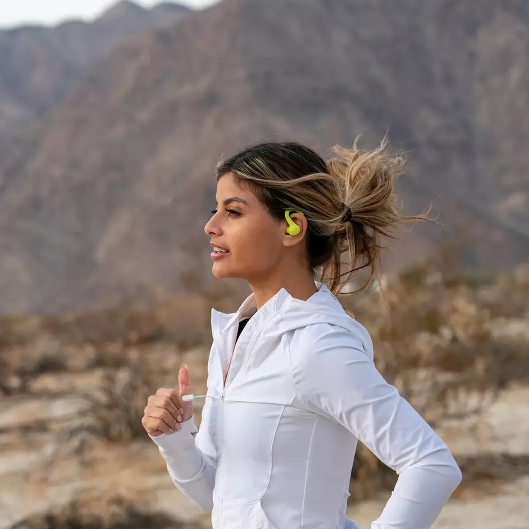 Kobieta biegnie na zewnątrz. Ma na sobie białą kurtkę, żółty słuchawki i włosy spięte w kok. Tłem jest pustynia.