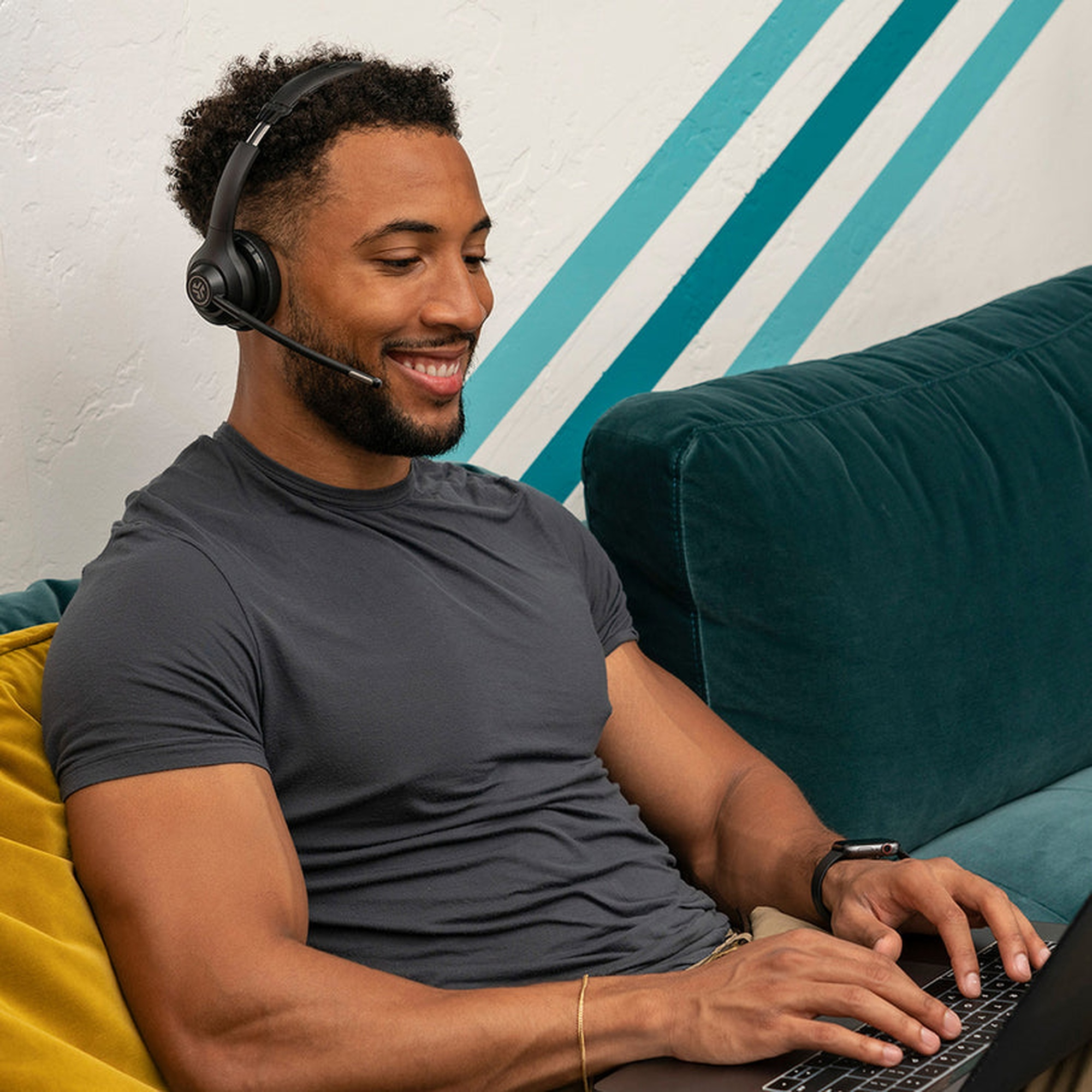 Mężczyzna w słuchawkach, uśmiechnięty i pracujący na laptopie. Siedzi na kanapie.