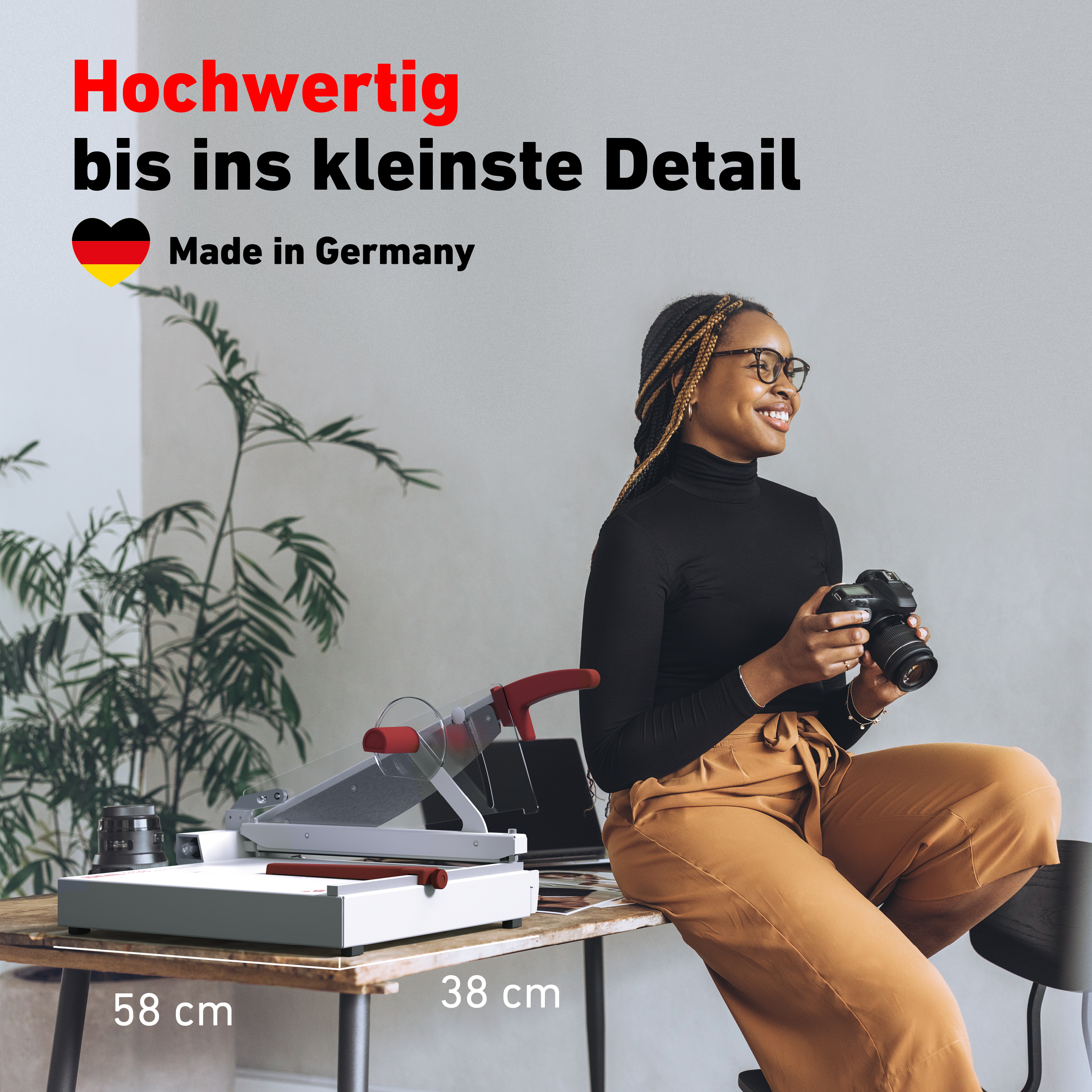 Frau mit Kamera, sitzend, neben Schneidemaschine auf Tisch. Made in Germany Text.