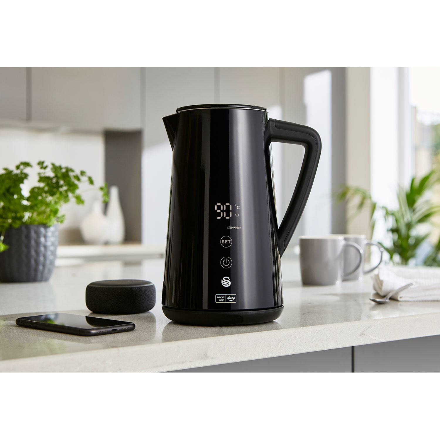 Schwarzer Wasserkocher mit Digitalanzeige, Smartphone und Smart Speaker auf Theke.