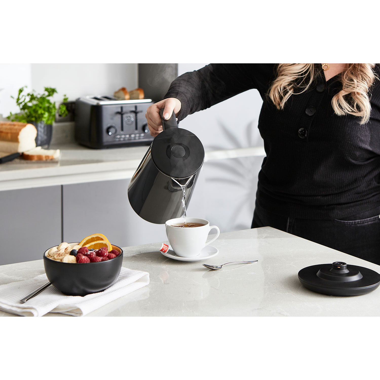 Eine Person gießt Wasser aus einem schwarzen Wasserkocher in eine weiße Teetasse. Eine Obstschale und ein Toaster sind auch vorhanden.