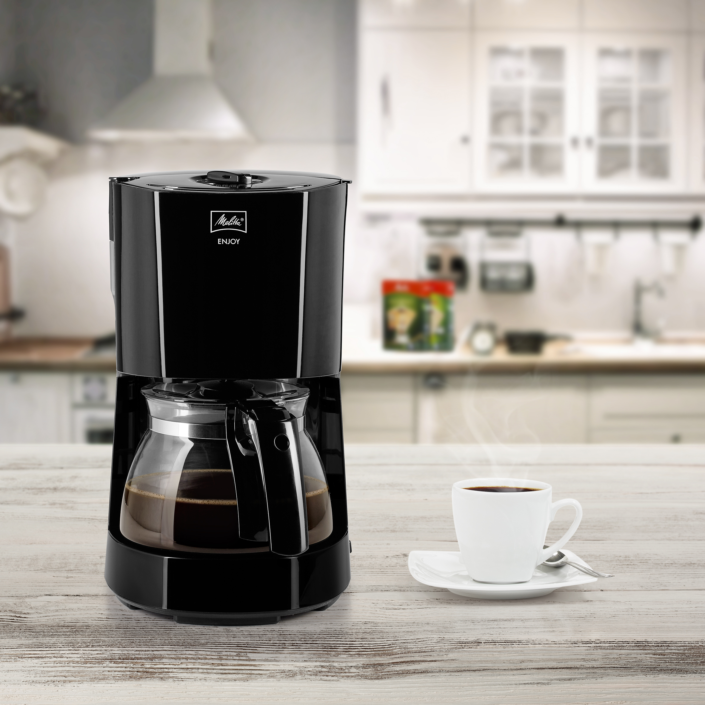 Czarny ekspres do kawy ze szklaną karafką, białą filiżanką z kawą i tłem kuchni.