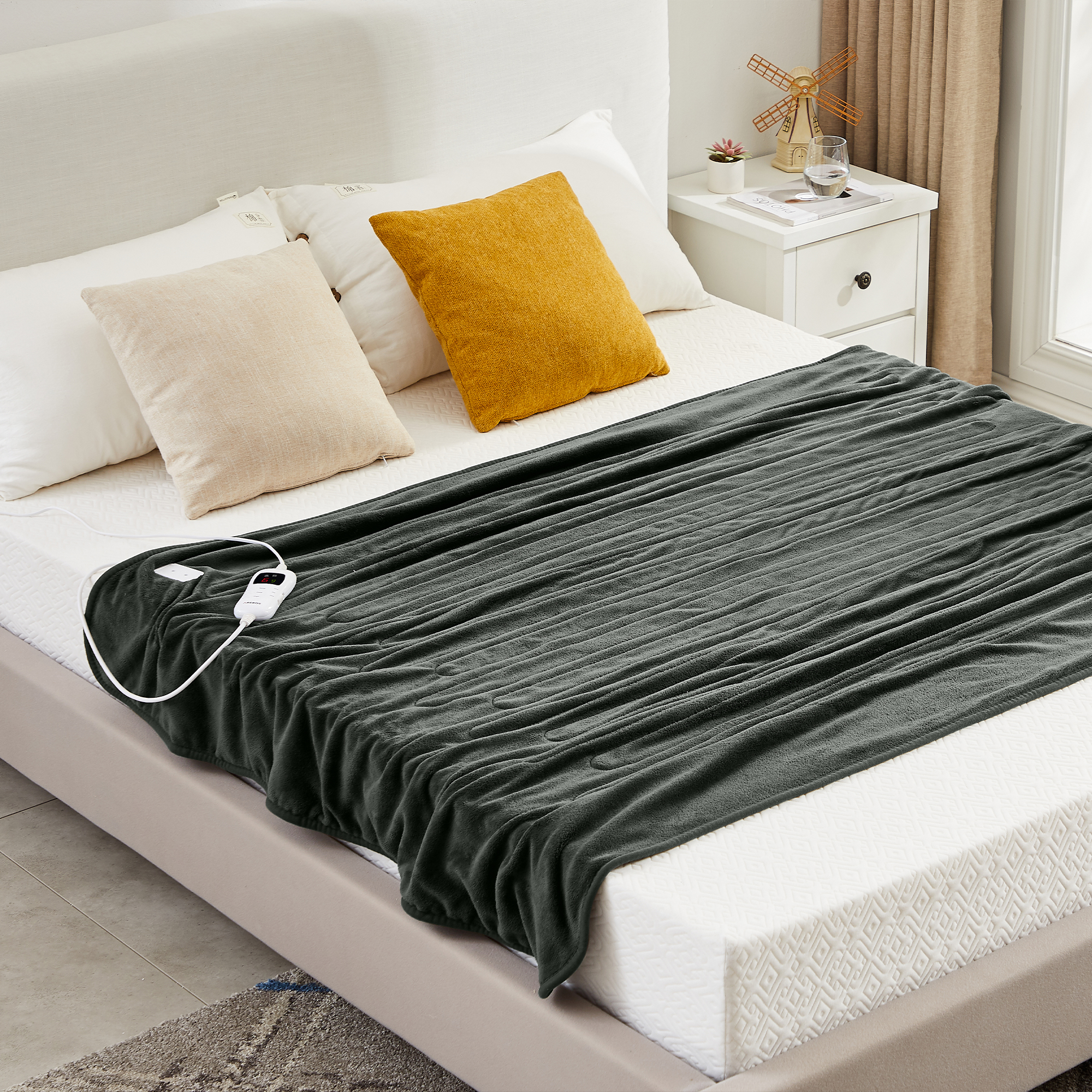 Dunkelgraue Heizdecke auf einem Bett mit weißen und gelben Kissen. Ein weißer Controller ist sichtbar.