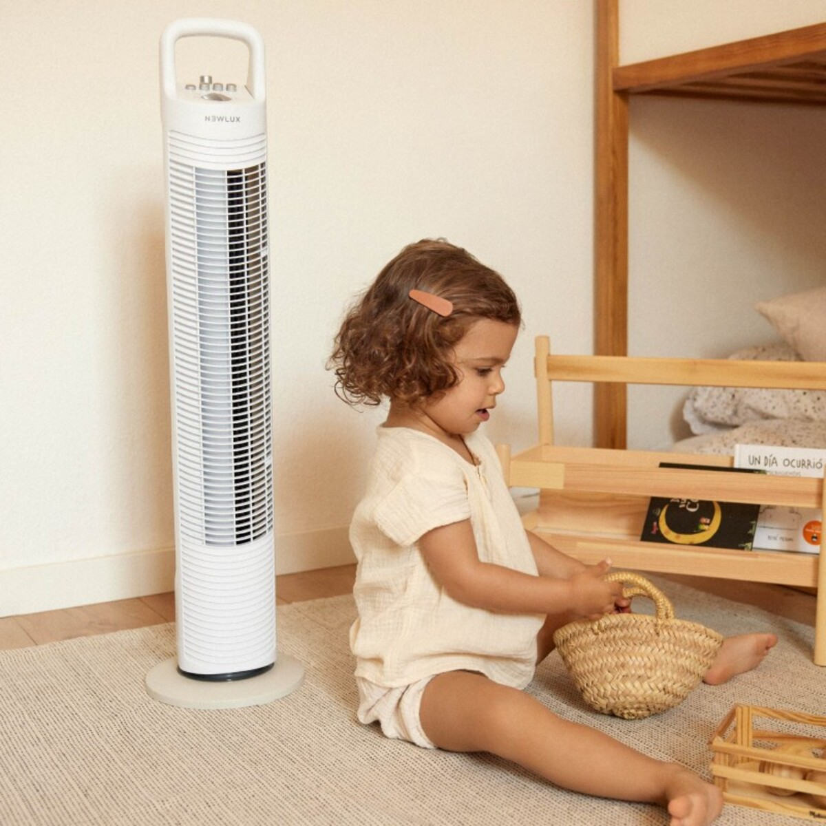 Un niño sentado cerca de un ventilador blanco. Una estantería de madera y una cesta de mimbre están cerca.