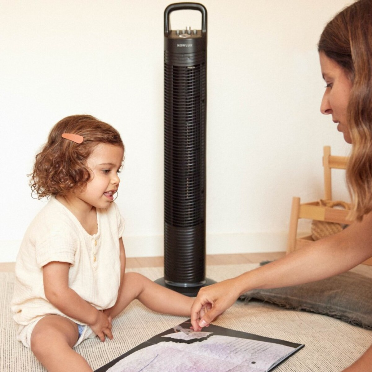 Un niño pequeño y una mujer mirando un dibujo. Un ventilador negro está detrás de ellos. Suelo blanco.