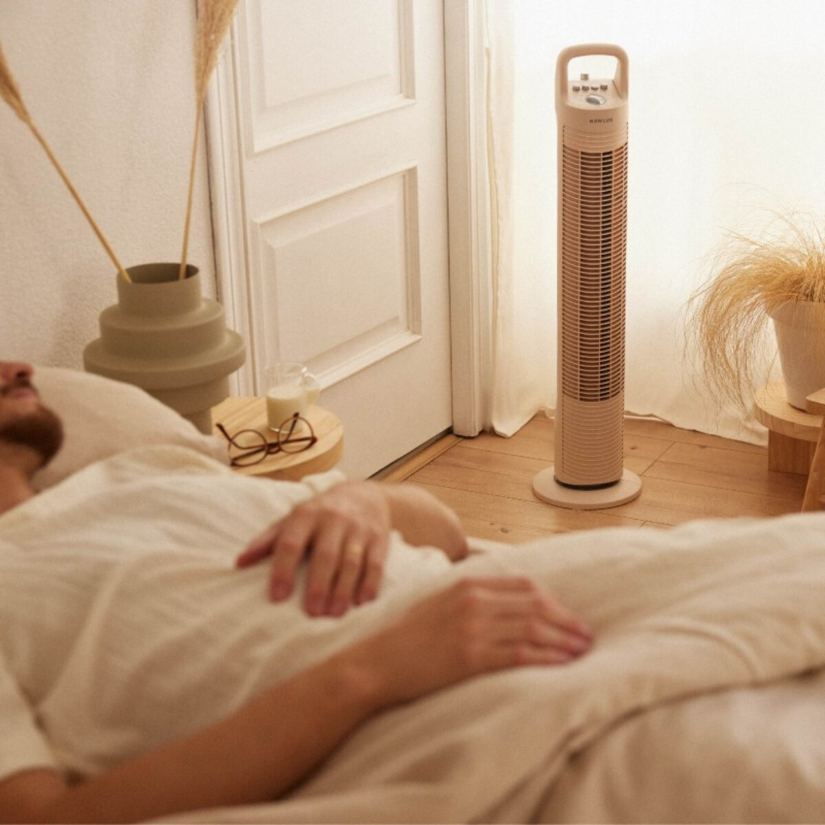 Escena de dormitorio con un hombre acostado en la cama, un ventilador y plantas en una mesa y un jarrón.