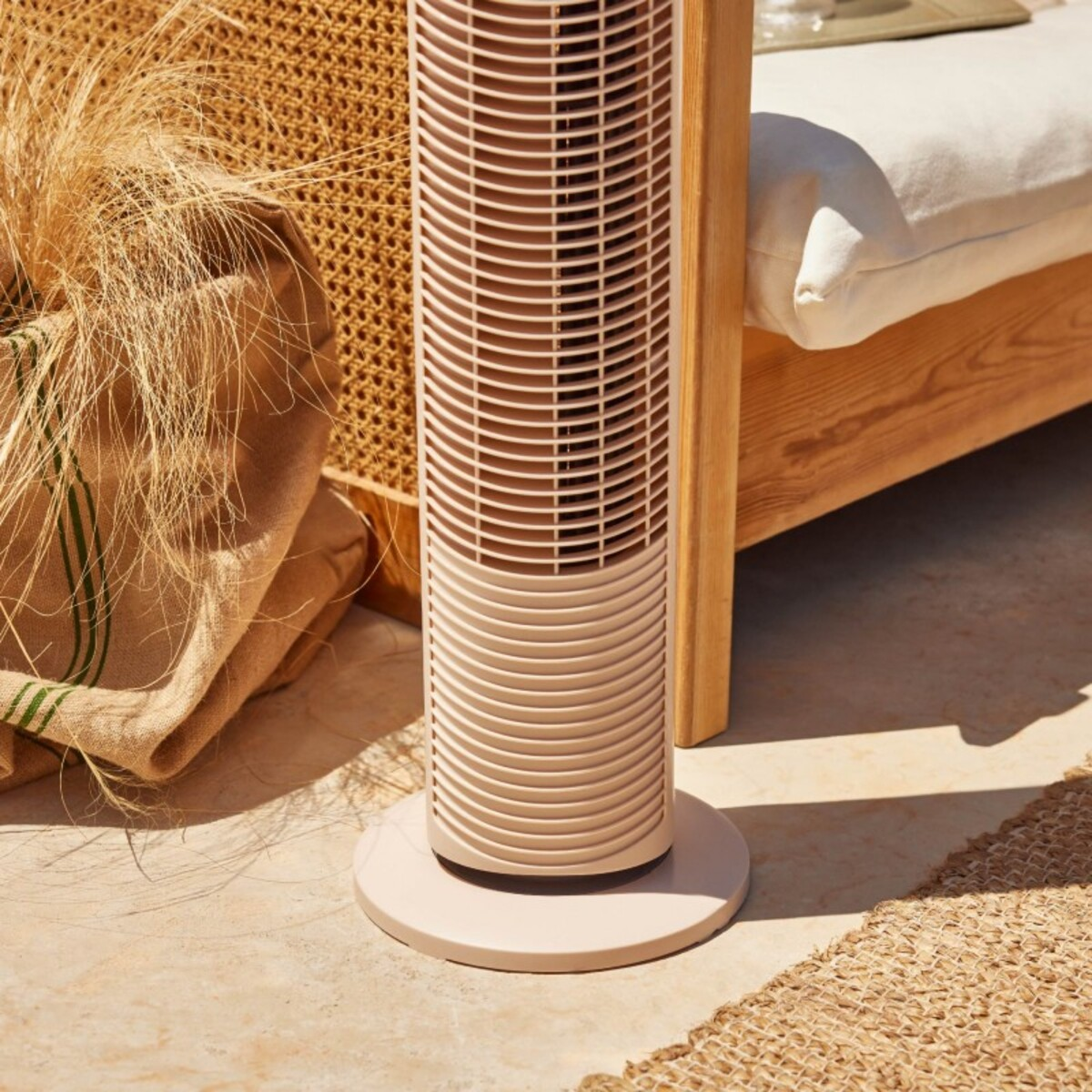 Un ventilador de torre beige sobre piso de baldosas, cerca de una silla tejida con un saco de arpillera.
