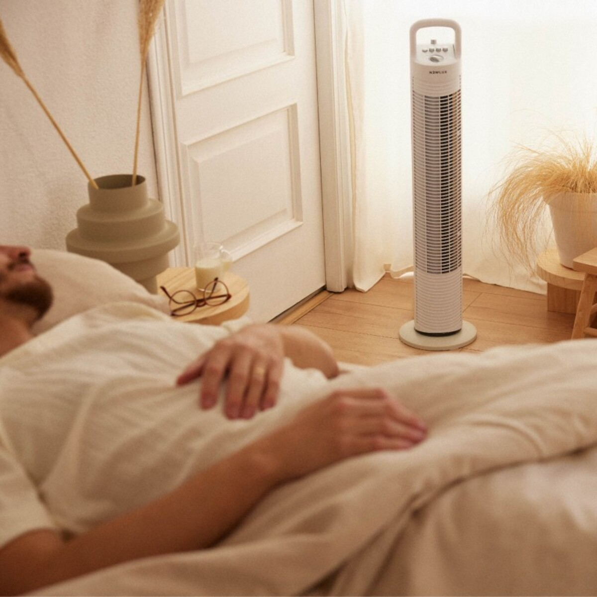 Hombre acostado en una cama con un ventilador de torre y una mesa con gafas. La habitación está bien iluminada.