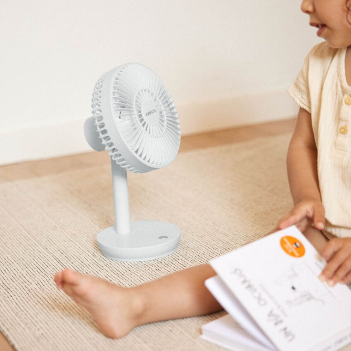 Niño con ventilador blanco y un libro sobre una alfombra.