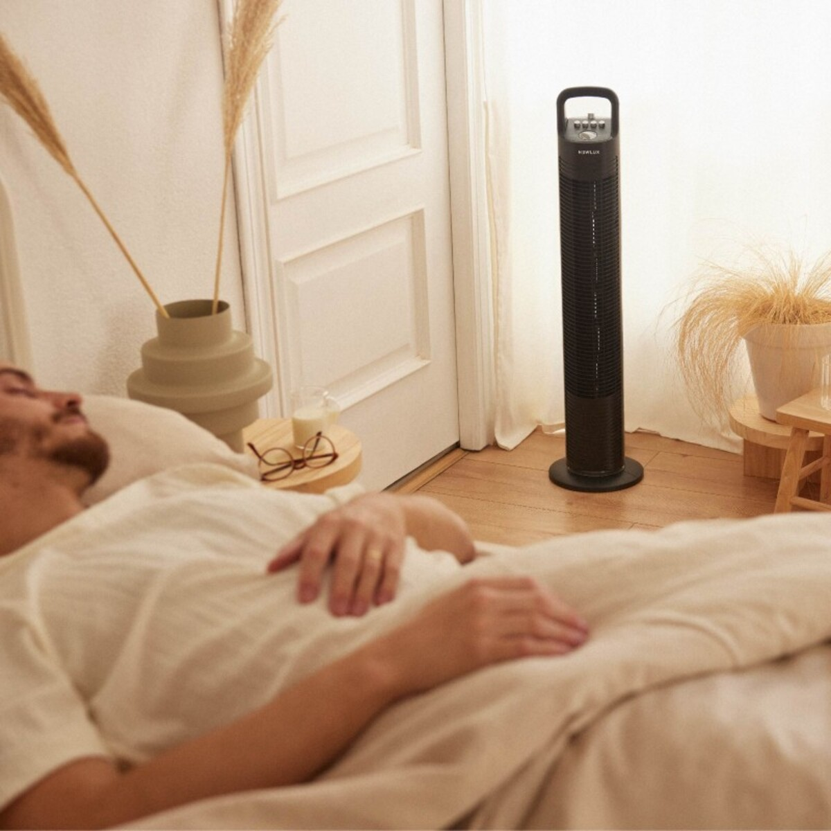 Un hombre durmiendo en la cama con un ventilador negro alto en la esquina. Plantas secas y gafas cerca.
