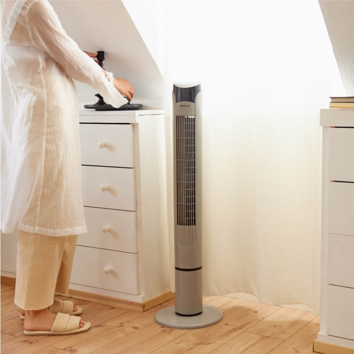 Una mujer con ropa blanca cerca de un ventilador alto gris. El ventilador está sobre una superficie blanca.