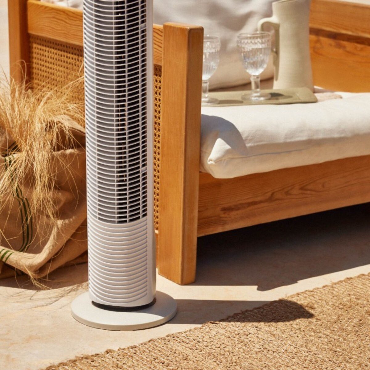 Un ventilador de torre blanco está junto a un banco de madera con cojines y una alfombra beige.
