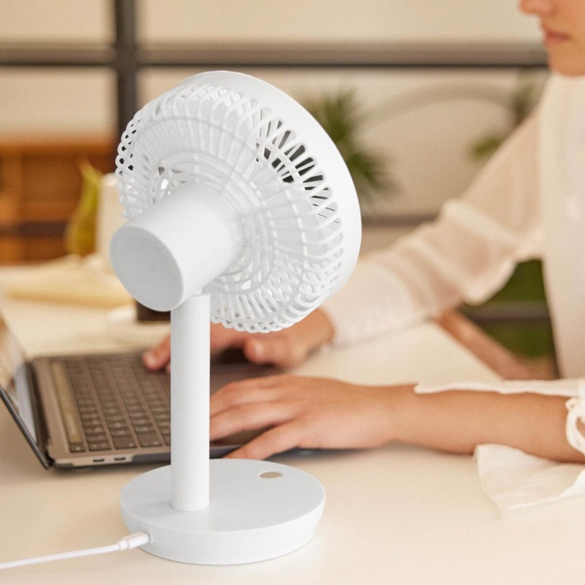 Ventilador de escritorio blanco sobre una base blanca. Persona trabajando en portátil.