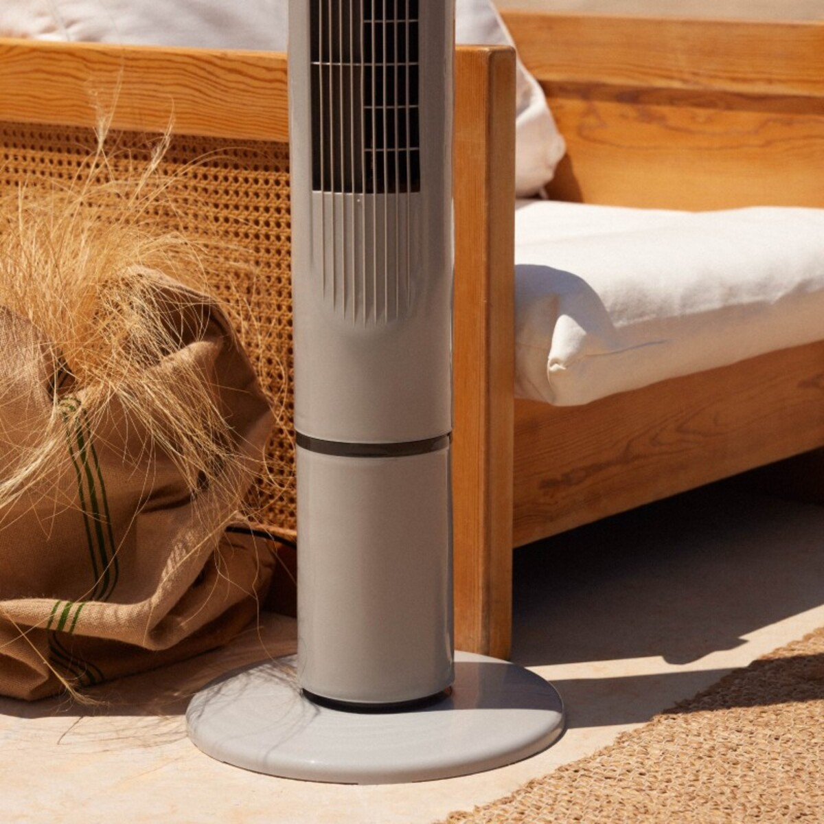 Un ventilador de torre gris sobre una base redonda, junto a una bolsa tejida y un marco de cama.