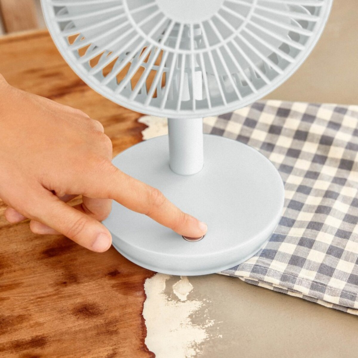 Una mano presiona el botón de encendido de un ventilador de escritorio azul claro sobre una mesa.