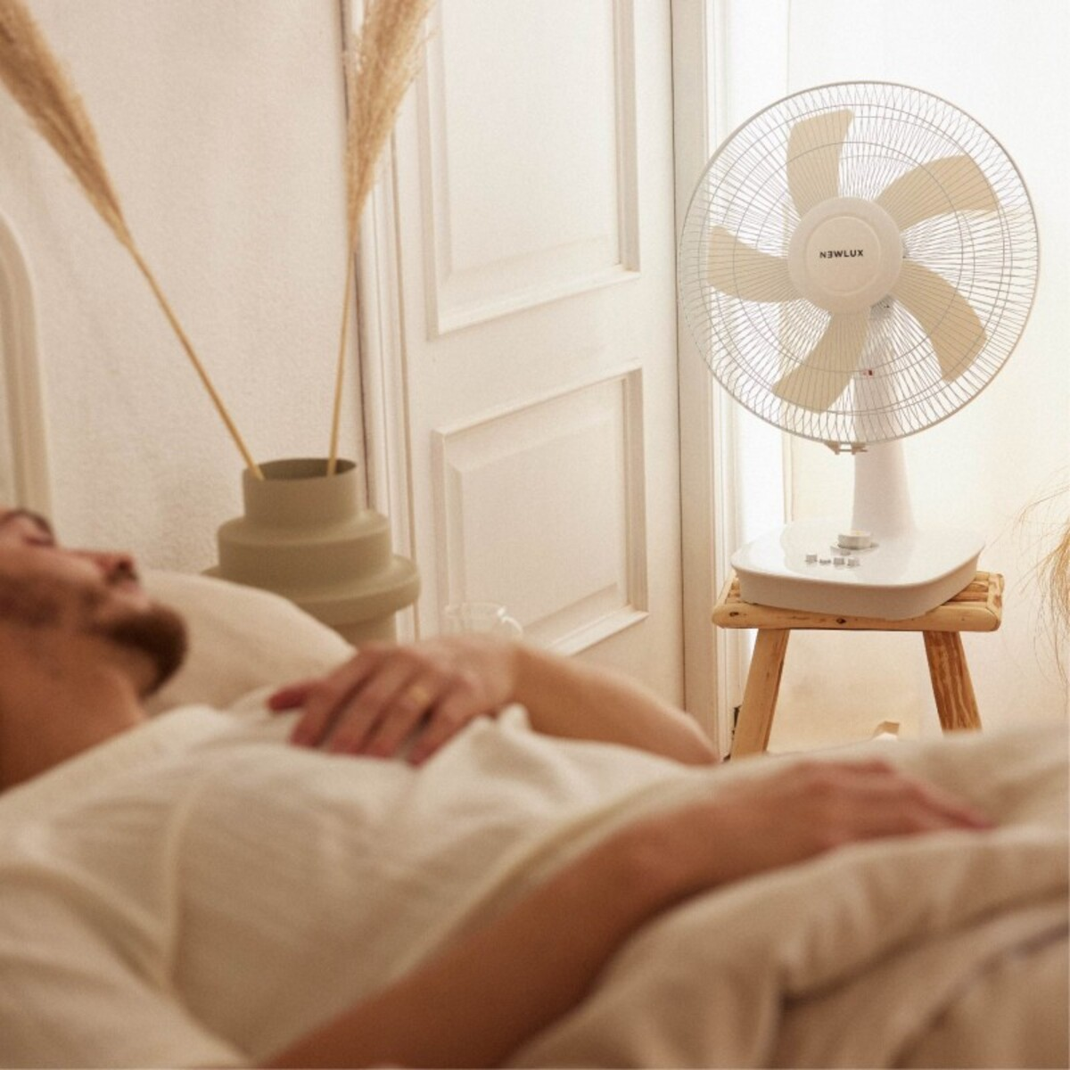 Hombre acostado en la cama con ventilador blanco en taburete.