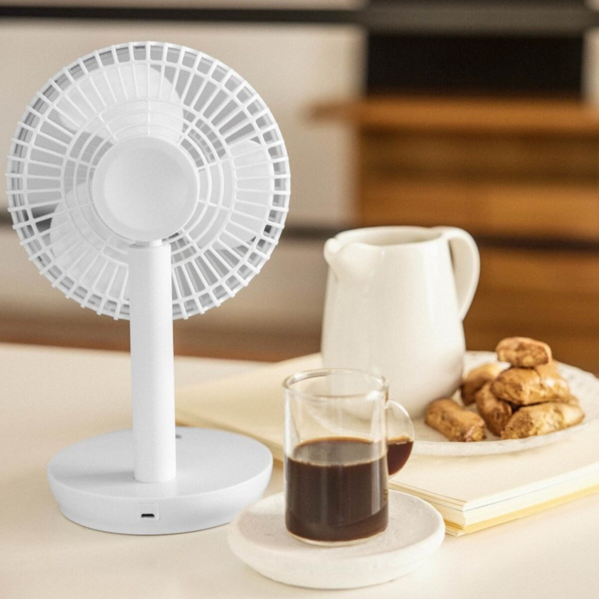 Ventilador de escritorio blanco, café en vaso, galletas y jarra. Ambiente brillante y limpio.