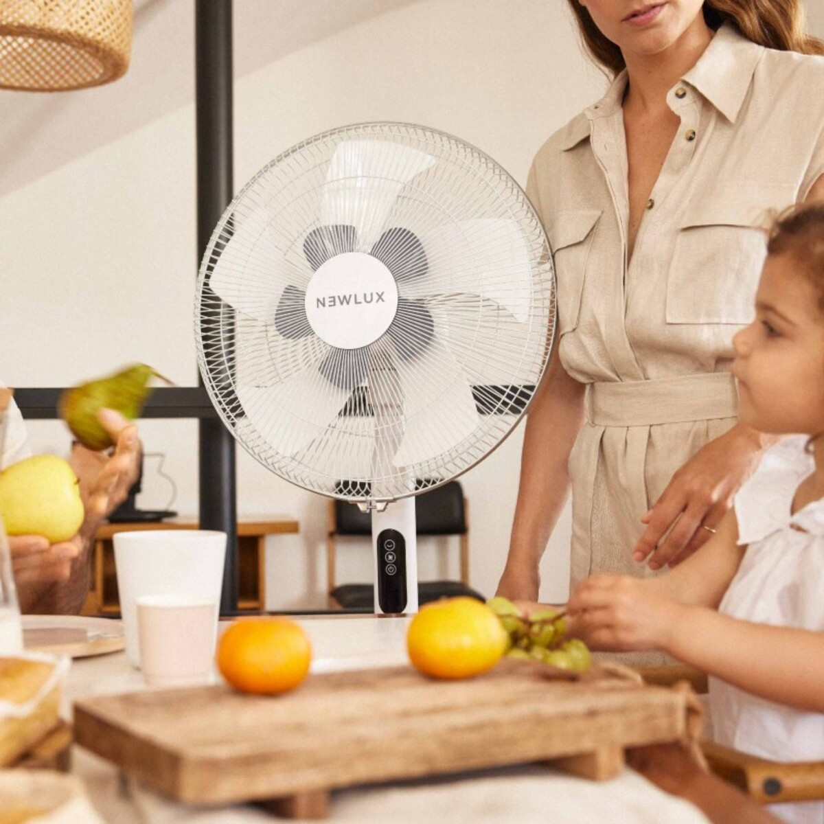 Un ventilador en una mesa con fruta y personas al fondo, una mujer está cerca.
