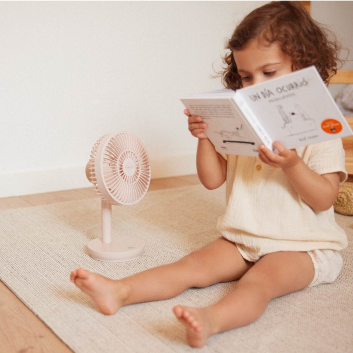 Niña leyendo un libro con un ventilador cerca; escena en una alfombra clara.