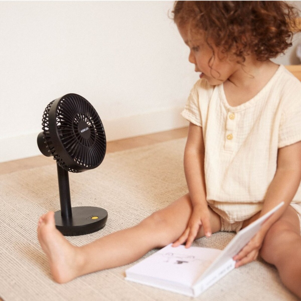 Una niña pequeña sentada en el suelo leyendo un libro mientras un ventilador negro le echa aire.