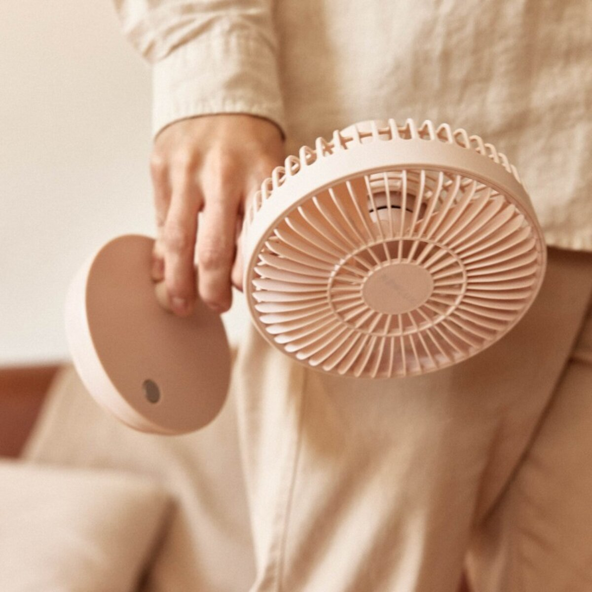Mano sosteniendo un ventilador portátil beige con soporte y base redonda. Ropa beige.