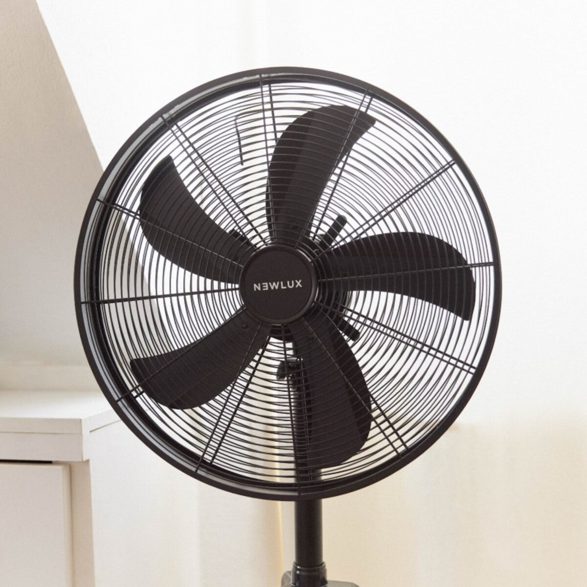 Ventilador de pie Newlux negro con cinco aspas y una jaula protectora. Fondo de pared blanca.