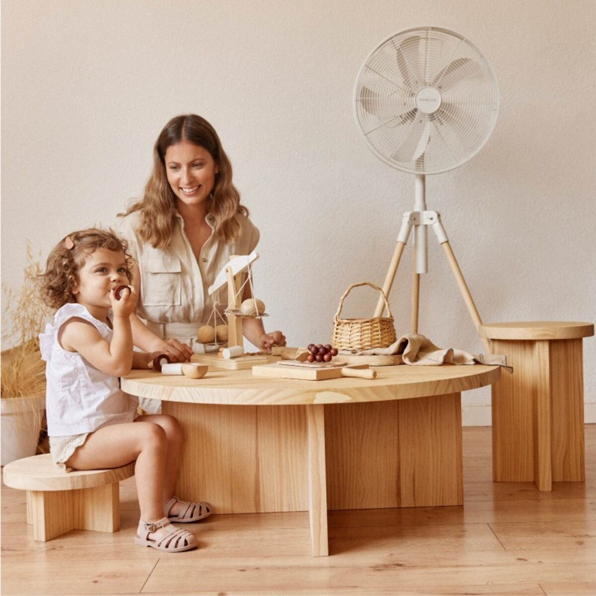 Una mujer y una niña comen en una mesa de madera con juguetes y un ventilador. Fondo blanco.