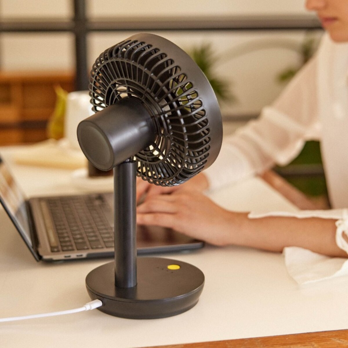 Ventilador de escritorio negro sobre una superficie blanca. Se ve el brazo de una persona. Cable blanco.