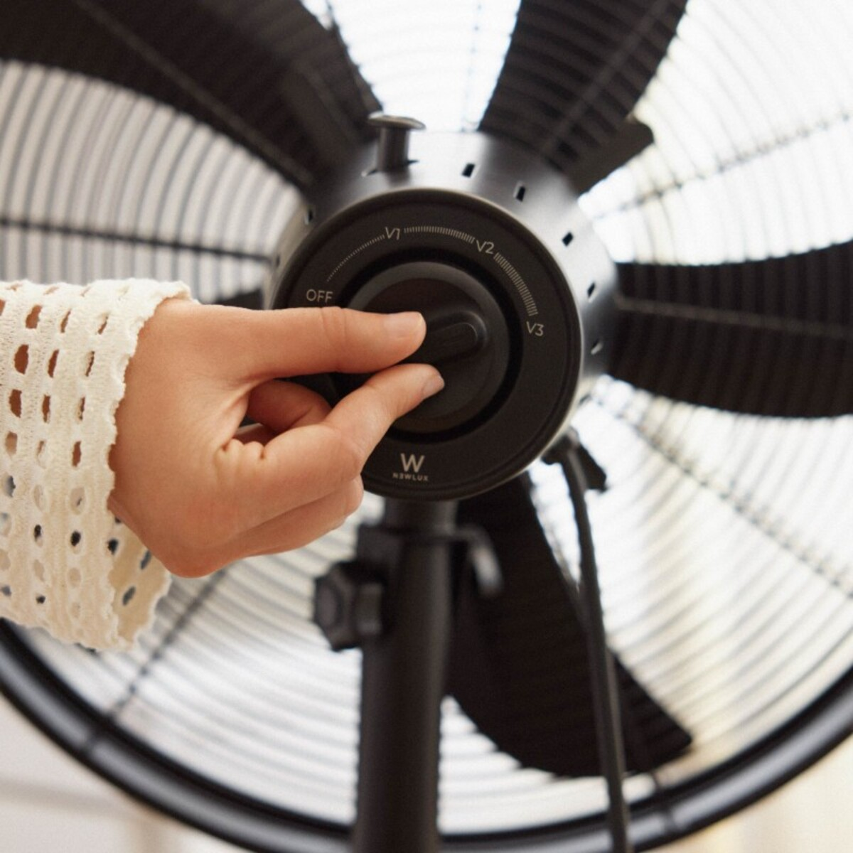 Una mano gira el dial de velocidad del ventilador. El ventilador es negro, con aspas visibles, y el dial muestra los ajustes de velocidad.