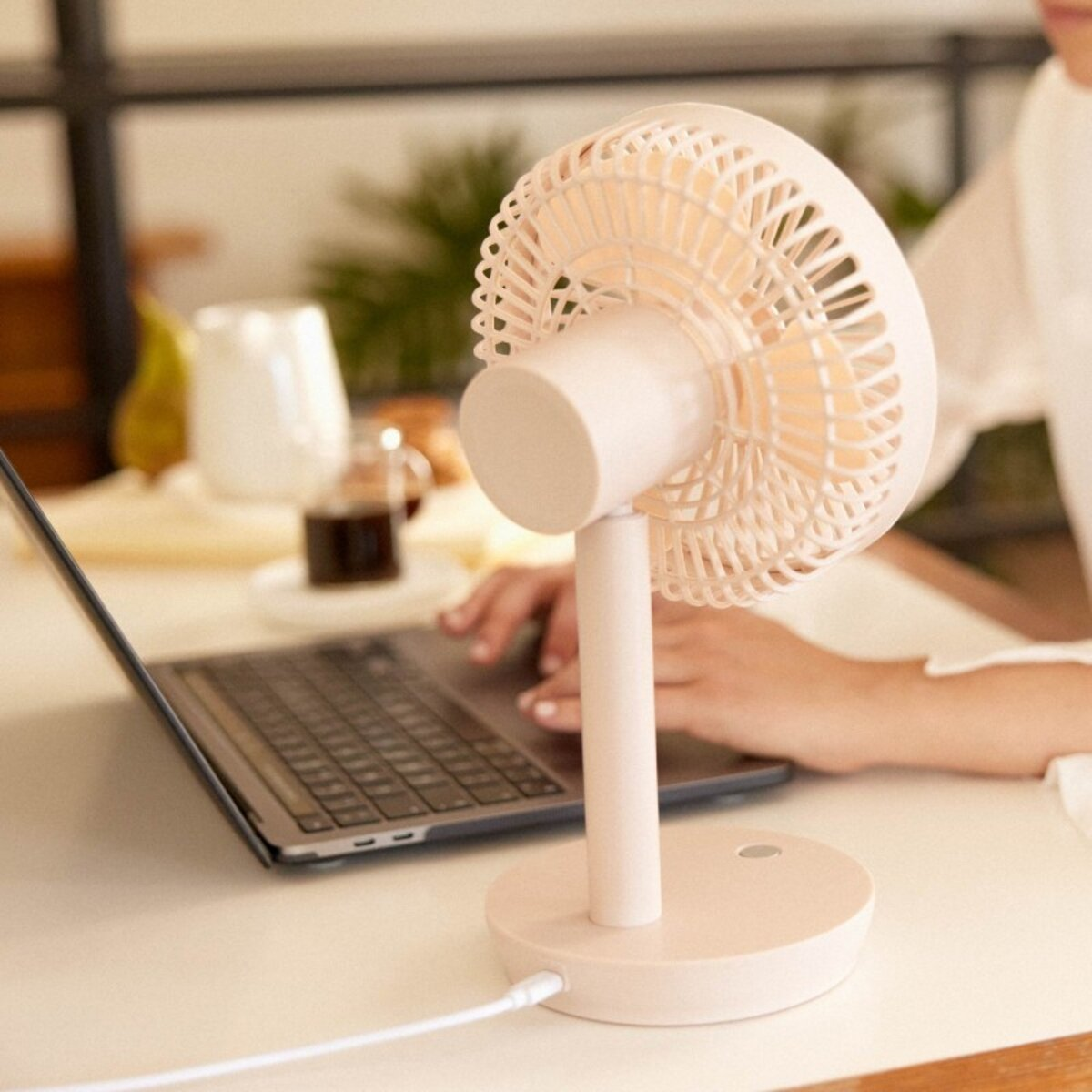 Ventilador de escritorio rosa con portátil y café al fondo, mesa blanca.