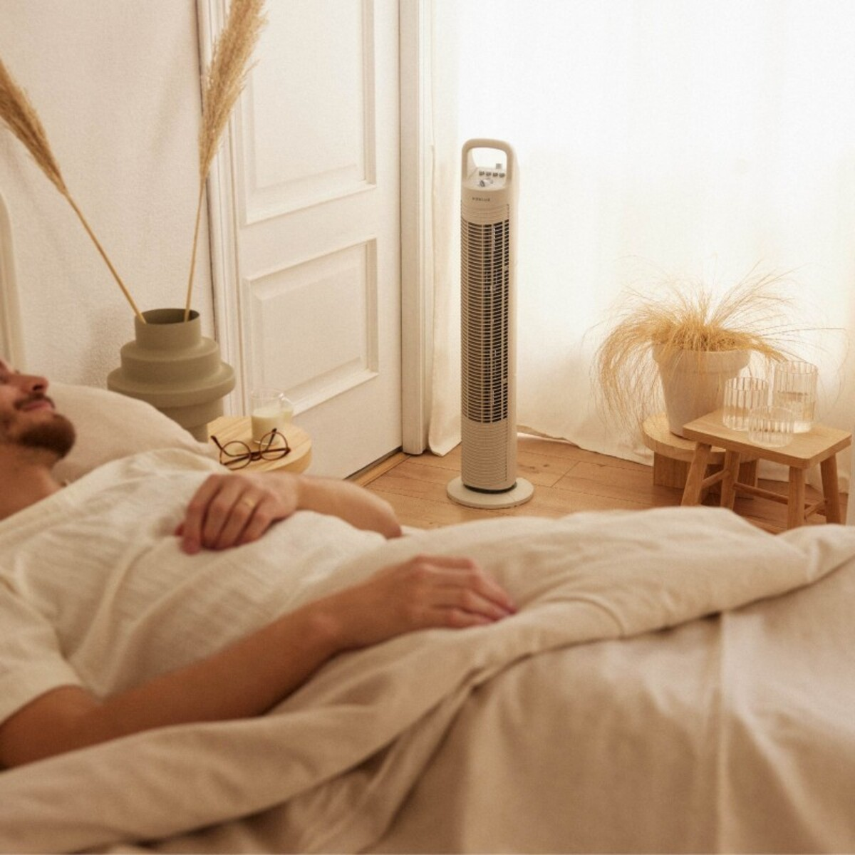 Un hombre está acostado en la cama junto a un ventilador y un jarrón. La habitación es beige y luminosa.