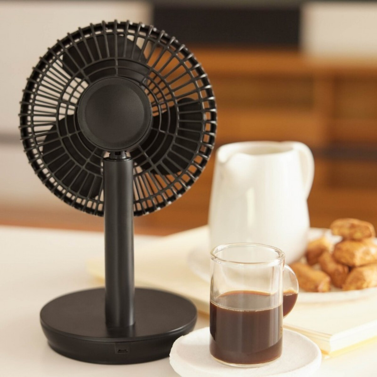 Ventilador de escritorio negro con aspas negras, sobre mesa blanca, con café y pasteles al fondo.
