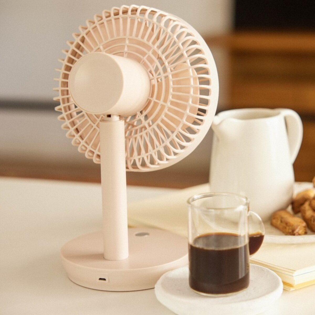 Un ventilador rosa, un vaso de café y una jarra blanca sobre una mesa.