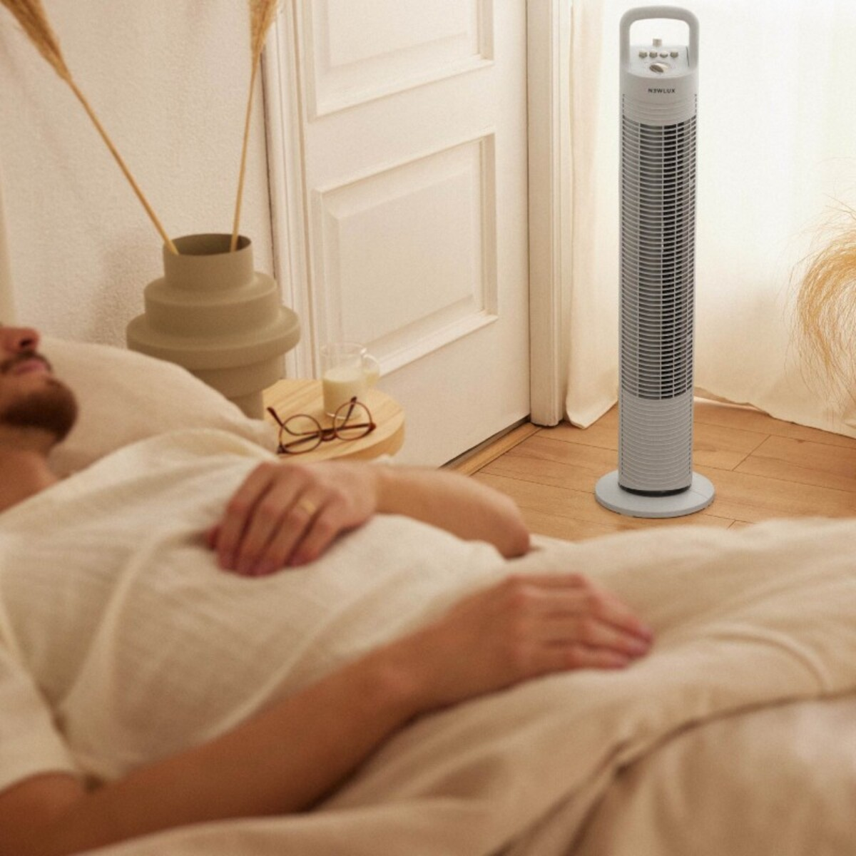 Hombre en cama, sábana blanca. Un ventilador y una mesita con gafas al fondo.