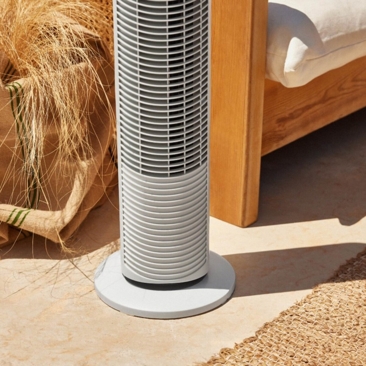Ventilador de torre blanco en suelo de piedra. Saco de paja y banco de madera al fondo.