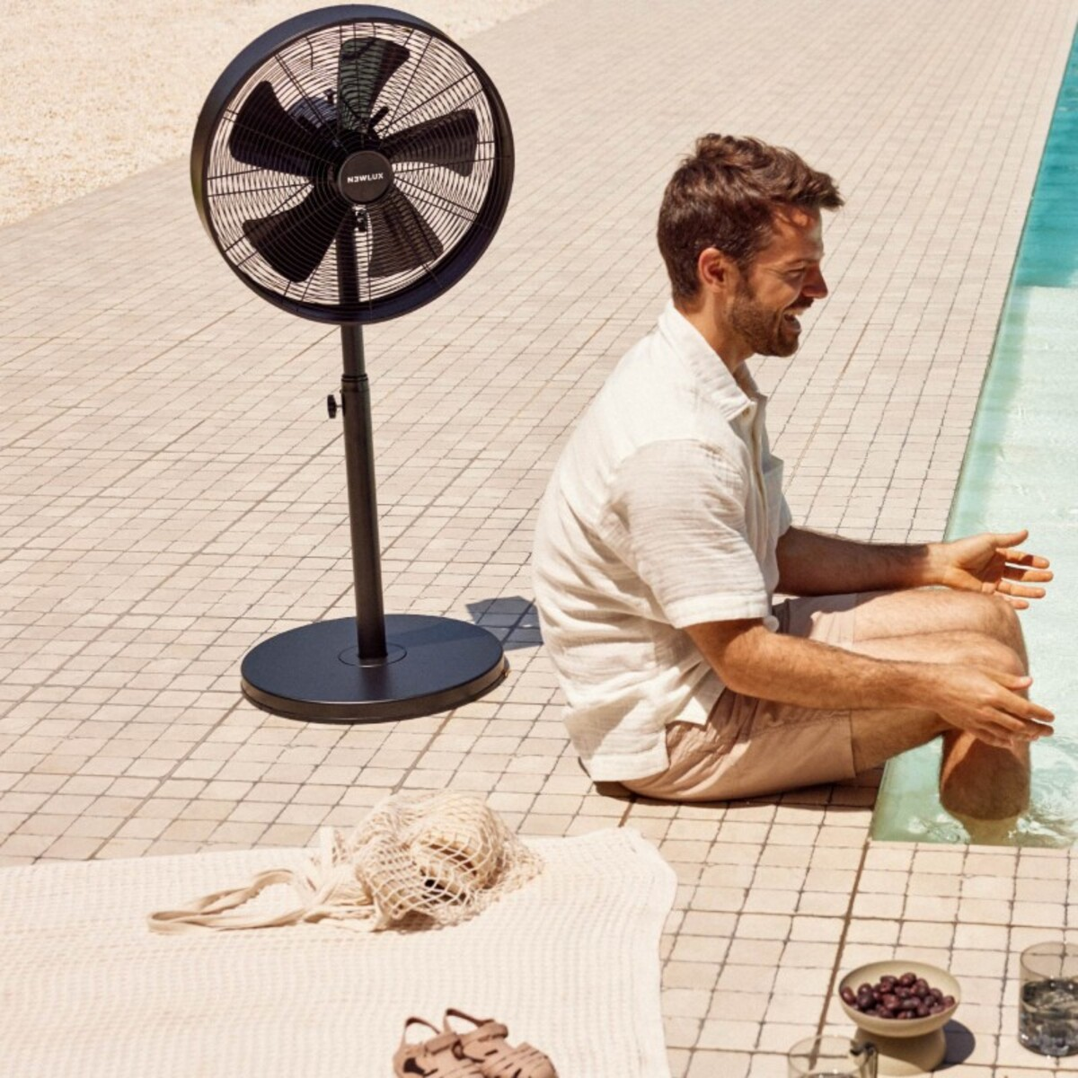Hombre sentado cerca de una piscina, ventilador soplando aire. Ventilador, agua y entorno soleado al aire libre.