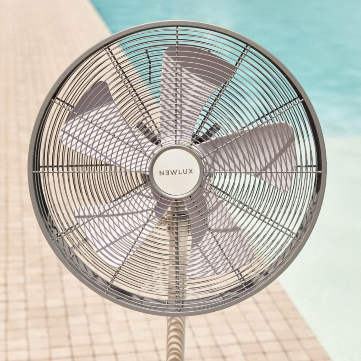 Ventilador Newlux gris con rejilla circular, junto a una piscina. El fondo es una piscina turquesa.