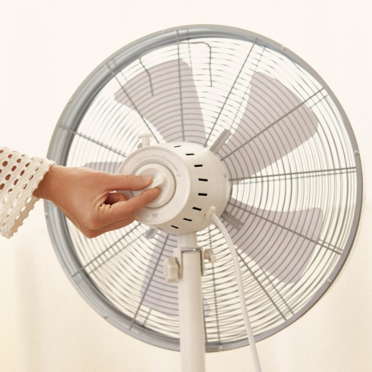Mano ajustando un ventilador blanco con jaula plateada, frente a una pared.