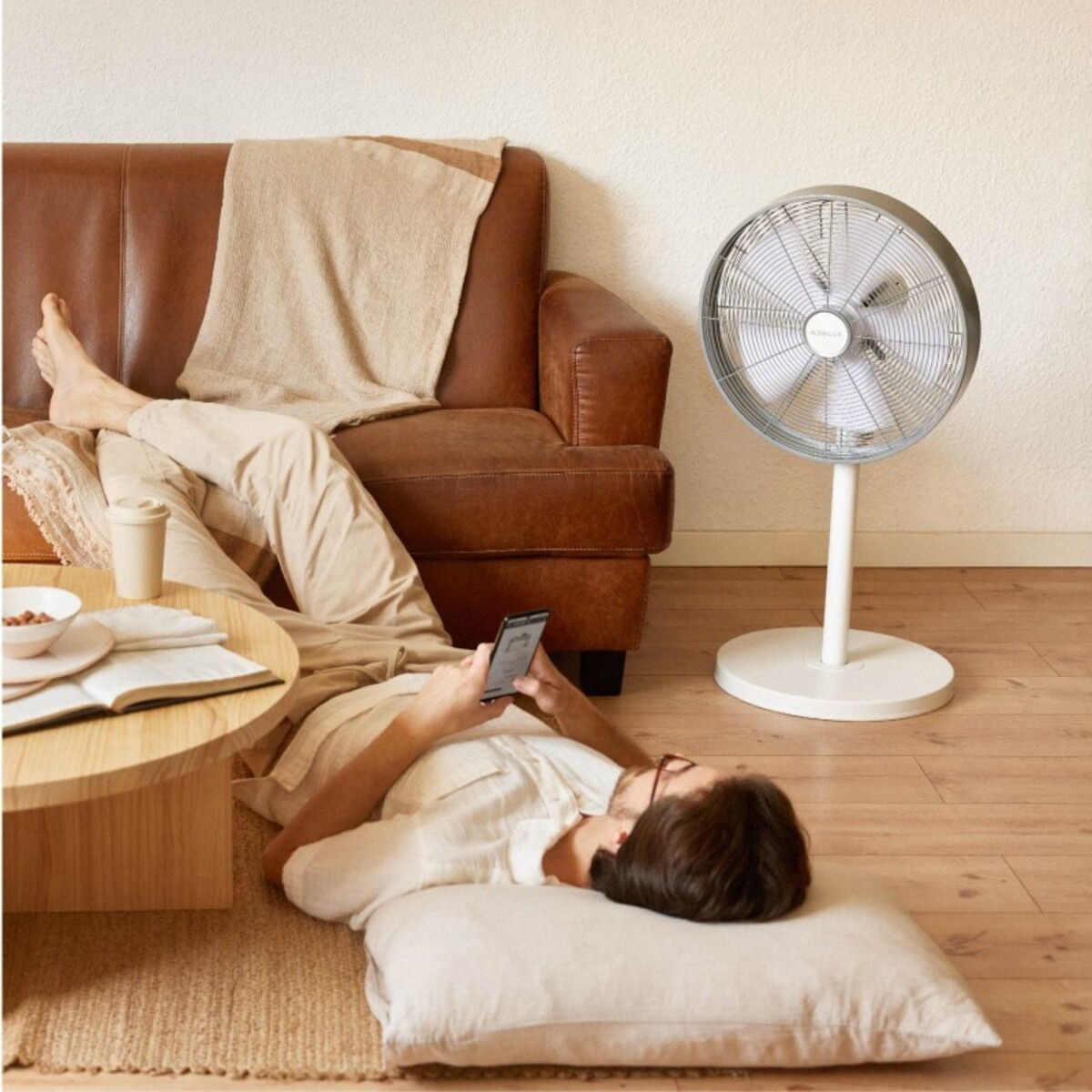 Hombre tumbado en el suelo, usando el teléfono. Ventilador a la derecha, sofá de cuero marrón. Ambiente acogedor.