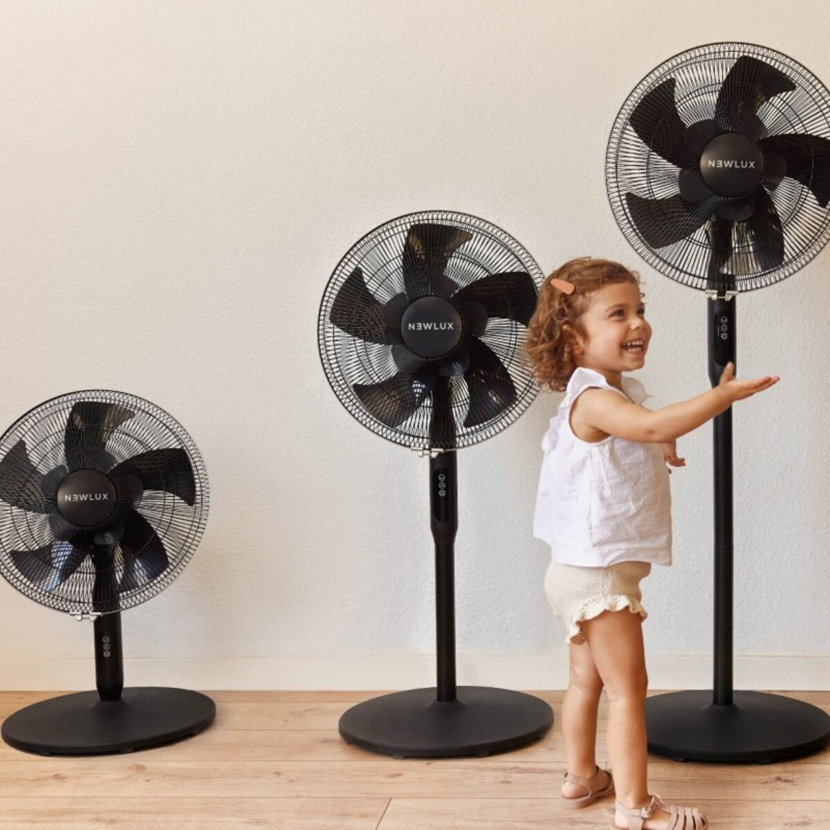 Una niña de pie cerca de tres ventiladores negros Newlux.