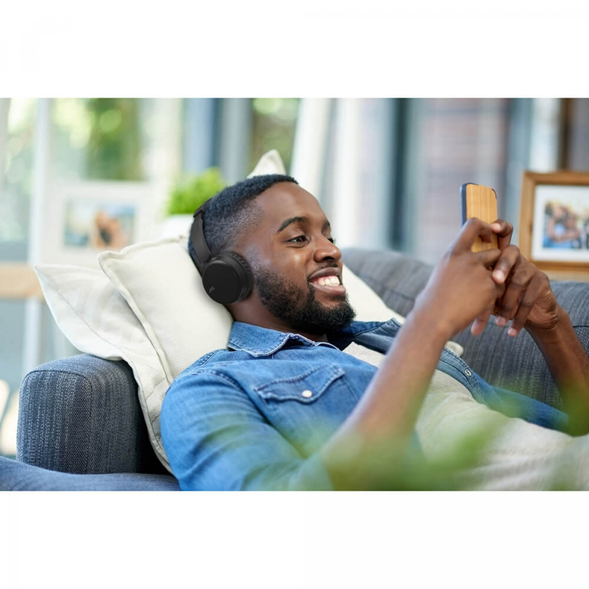 Mężczyzna leżący na sofie w słuchawkach i patrzący na smartfon, uśmiechnięty.