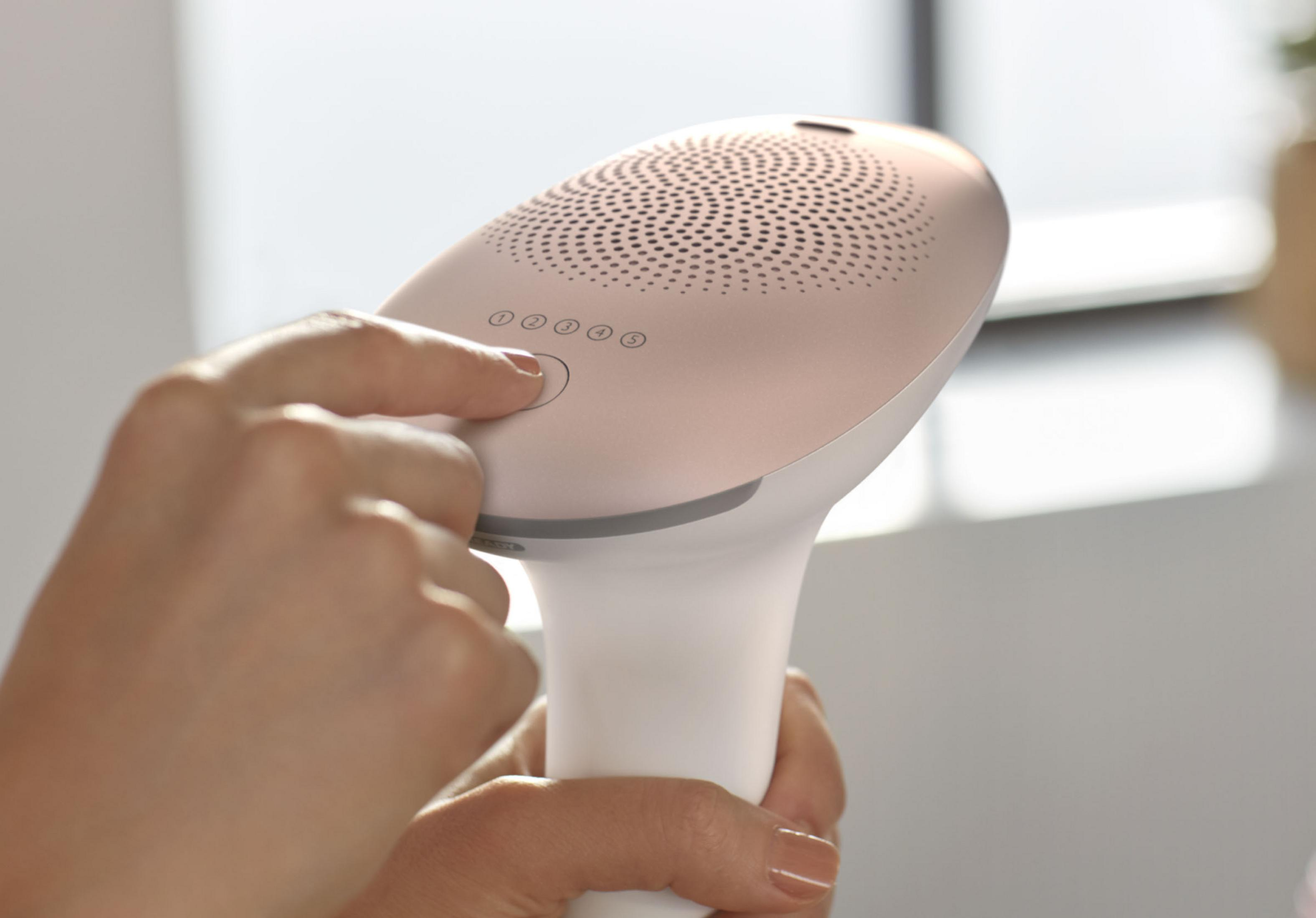 Persona che preme un pulsante su un dispositivo di epilazione rosa e bianco. Sfondo bianco.