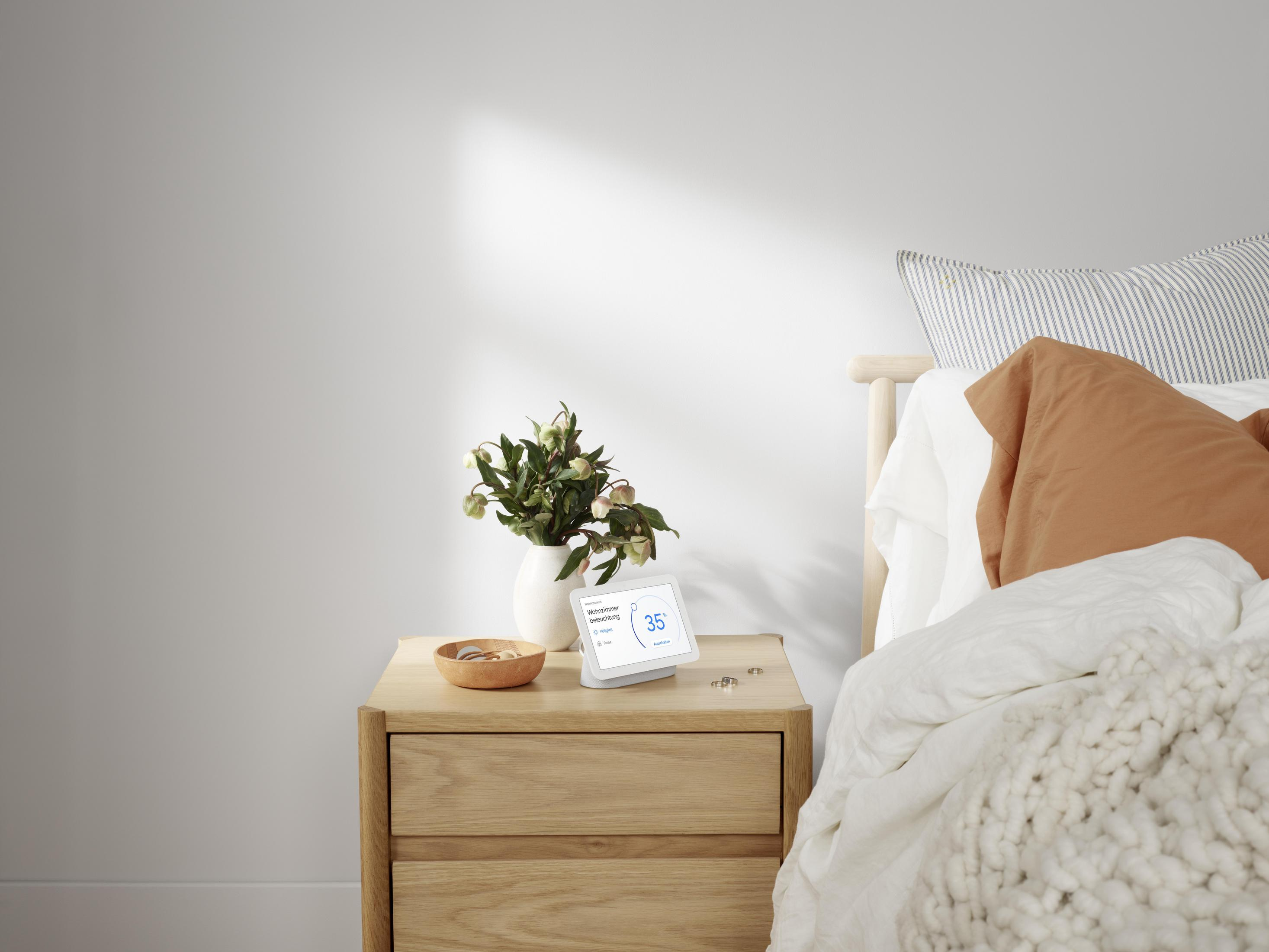 Display intelligente bianco su comodino con vaso, fiori e letto.