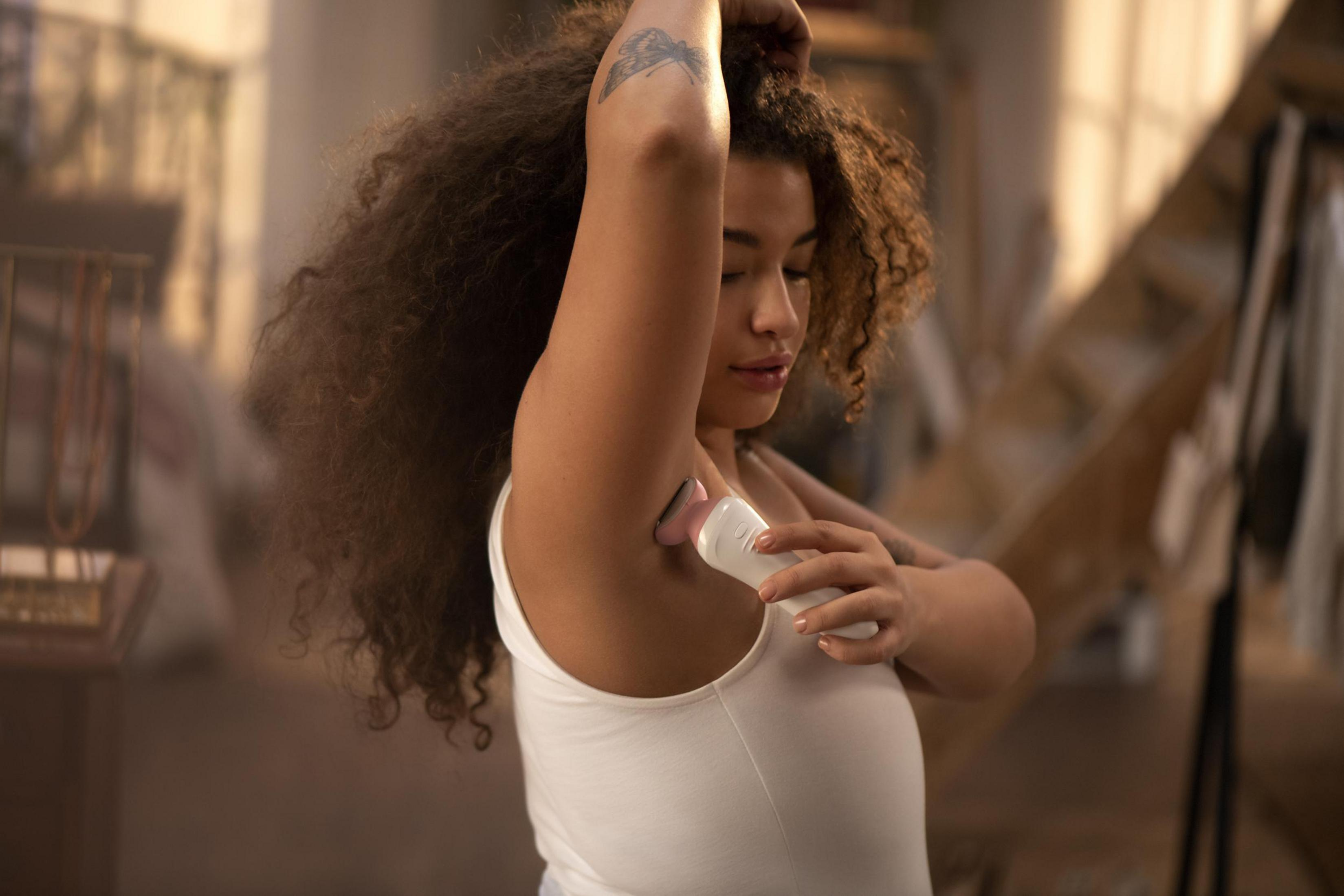 Kobieta używa depilatora pod pachą, ramiona w górze, w miękko oświetlonym pomieszczeniu.