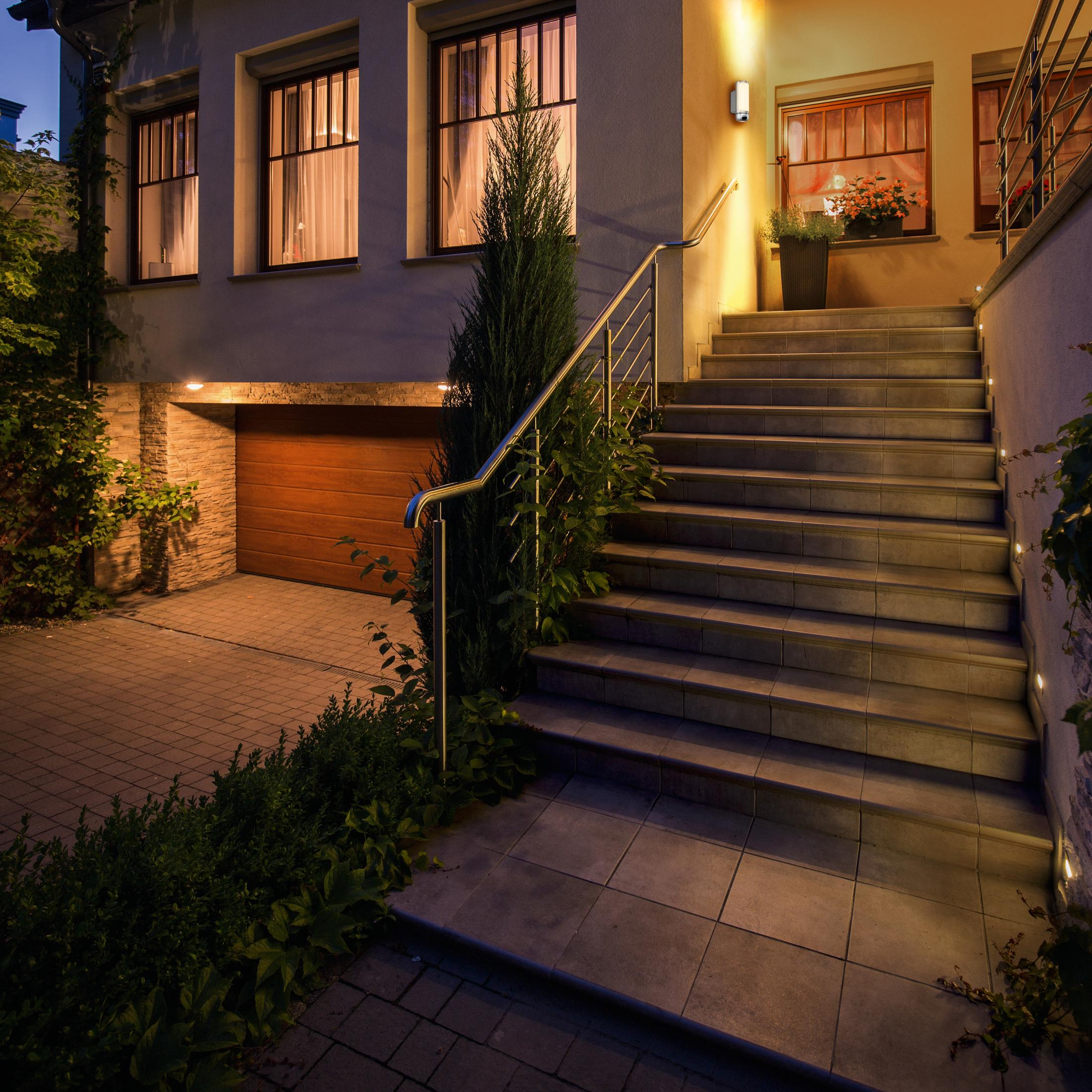 Entrada iluminada de una casa por la noche. Escaleras conducen a la puerta, con garaje y follaje.