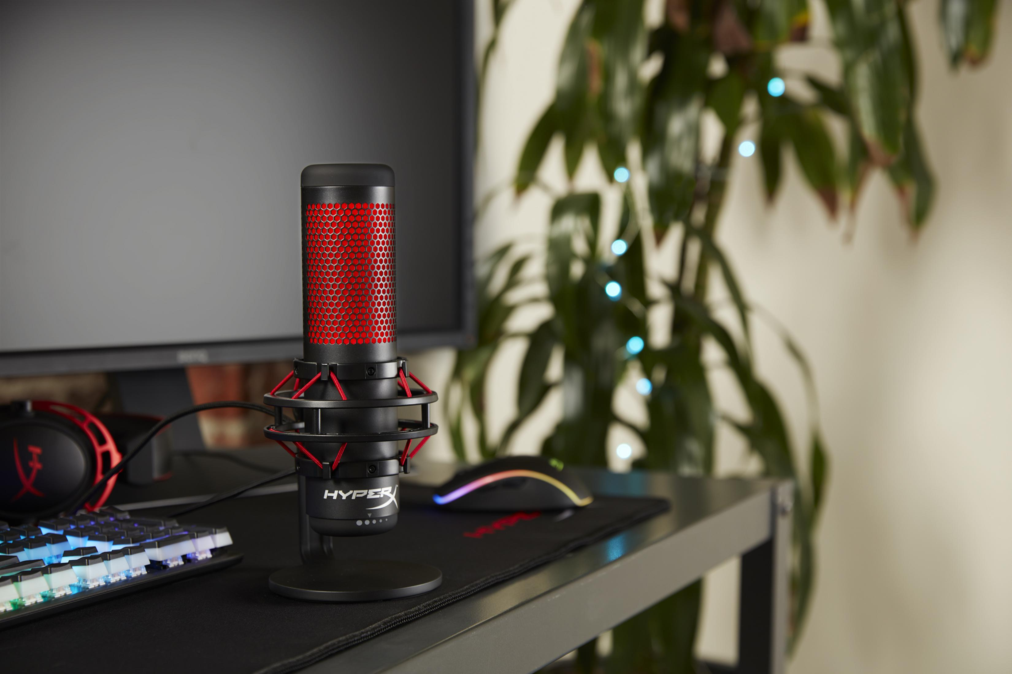 Czerwony i czarny mikrofon HyperX stoi na biurku z monitorem i klawiaturą w tle.